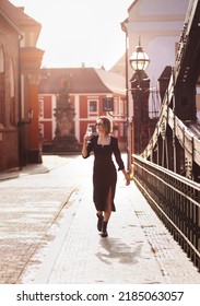 Attractive Happy Girl In A Black Dress  Walks Along A City Street With A Glass Of Wine. Glamorous Lifestyle. Fashion.