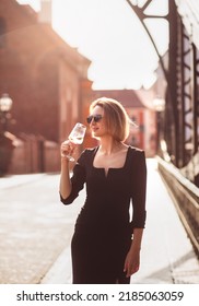 Attractive Happy Girl In A Black Dress  Walks Along A City Street With A Glass Of Wine. Glamorous Lifestyle. Fashion.