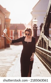 Attractive Happy Girl In A Black Dress  Walks Along A City Street With A Glass Of Wine. Glamorous Lifestyle. Fashion.