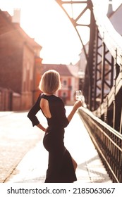 Attractive Happy Girl In A Black Dress  Walks Along A City Street With A Glass Of Wine. Glamorous Lifestyle. Fashion.