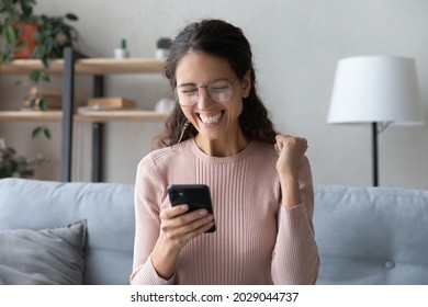 Attractive happy Latina woman in glasses sit on sofa at home holding smartphone read sms notification great news looking excited overjoyed cant believe in luck enjoy moment of victory. Success concept - Shutterstock ID 2029044737
