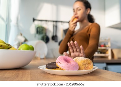 Attractive Happy Latino Woman Pick Up A Green Apple In Kitchen At Home. Young Beautiful Female Desire To Chose Fruits And Ignore Sweet Donut On Table, Enjoy Eat Healthy Foods For Health To Lose Weight