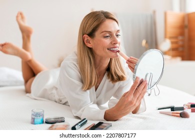 Attractive happy middle aged woman applying lipstick, looking at mirror while lying on bed and putting on decorative makeup, using cosmetic product indoors - Powered by Shutterstock