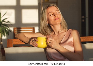 Attractive happy middle aged woman is sitting on  sofa in living room. Smiling adult lady enjoys drinking coffee or tea sitting on couch at home