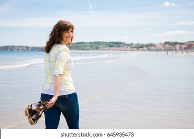 Attractive and happy middle aged woman walking along the seashore holding the shoes in hand