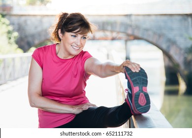 Attractive And Happy Middle Aged Woman Stretching Legs Using A Trail Outdoors