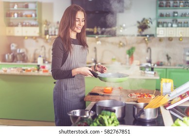 Attractive happy slim Caucasian female cooking pasta, making sauce using a pan at home.