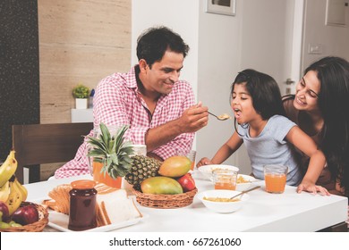 An Attractive Happy, Smiling Asian Indian Family Of Mother, Father And Daughter. Father Feeding Cereal To Daughter With Spoon At Dining Table. Indians Eating Breakfast, Lunch / Dinner. Selective Focus