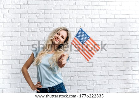 Attractive happy smiling caucasian girl posing with American and British flags, she stands near a white brick wall, smiling and looking at the camera