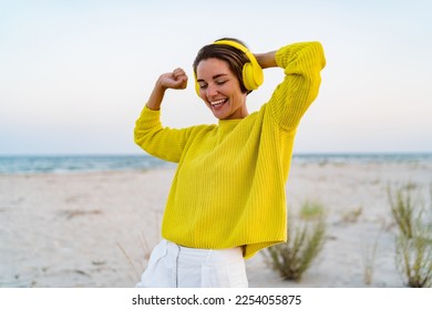 attractive happy smiling woman listening to music in colorful yellow headphones on beach in summer style fashion trend outfit happy, wearing yellow sweater having fun dancing singing