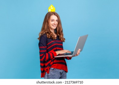 Attractive happy woman wearing striped casual style sweater, holding laptop in hands, looking at camera, holding paper house on head, online renting. Indoor studio shot isolated on blue background. - Shutterstock ID 2127369710