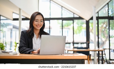 Attractive and happy young Asian businesswoman or female office worker in formal suit and eyeglasses working on her project assignment on laptop computer in cafe co-working space. - Powered by Shutterstock