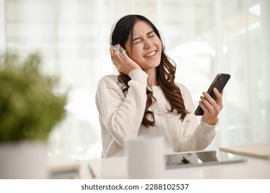 Attractive and happy young Asian woman in casual clothes wearing headphones, relaxes listening to music in her home office. - Shutterstock ID 2288102537