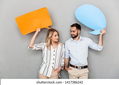 Attractive happy young couple standing isolated over gray background, holding empty speech bubbles