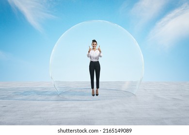 Attractive happy young european businesswoman showing thumbs up under and inside big glass sphere cover on concrete ground and bright blue sky background. Protection and limitation concept