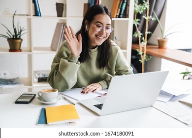 Attraktive, glückliche junge Studentin, die in der Universitätsbibliothek studiert, am Schreibtisch sitzend, mit Laptop-Computer, Videochat, winkend