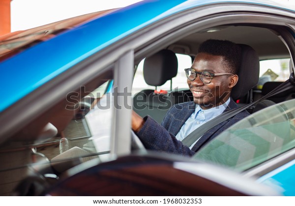 Attractive happy young man driving
car and smiling. Young man is choosing a new vehicle in car
dealership. Smiling handsome African American man driving his
car