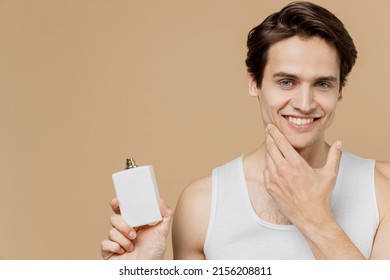 Attractive happy young man he 20s perfect skin wear undershirt hold aftershave deodorant bottle touch chin isolated on plain pastel beige background. Skin care healthcare cosmetic procedures concept.