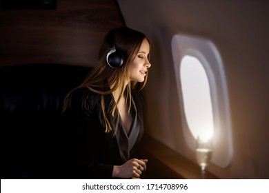 Attractive happy young woman with long blonde hair, in black jacket dress, sitting in an airplane seat with closed eyes, listening to music with headphones, enjoying the flight.