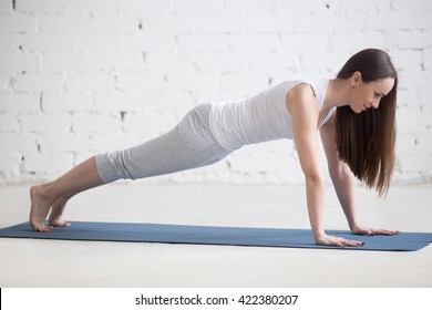Attractive happy young woman working out indoors. Side view portrait of beautiful model doing exercises on blue mat. Standing in plank pose (phalankasana). Full length