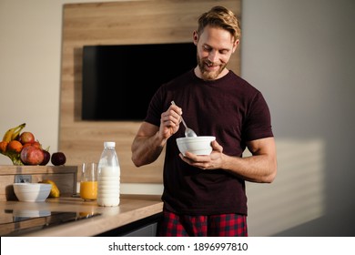 Attractive healthy young man having tasty breakfast while standing in the kitchen, holding bowl - Powered by Shutterstock