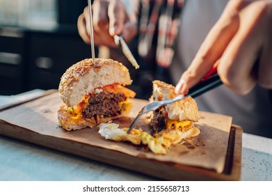 Attractive Hipster Dressed In Checkered Shirt Eating A Vegan Burger In A Restaurant. High Calorie Food. Cheat Meal. Focus On A Delicious Burger On A Wooden Plate.