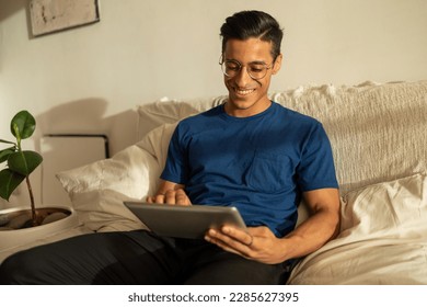 Attractive Hispanic man with glasses smiling wearing blue shirt siting on the sofa in the apartment using tablet  - Powered by Shutterstock