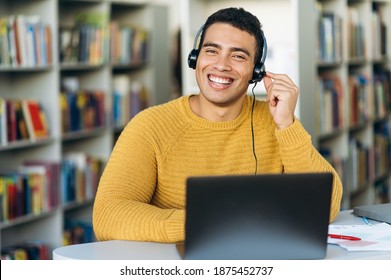 Attractive Hispanic Young Man, Call Center Worker, Student, Or Successful Business Man, In A Headset Is Using A Laptop While Working Online Sitting At His Desk And Looking At The Camera With Smile