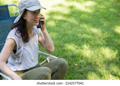 Attractive Hispanic Young Woman Wearing A Cap Talking On Mobile Phone And Doing Home Office While Sitting In A Lounge Chair At The Backyard In The Springtime. Copy Space.