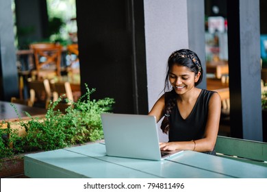 attractive indian asian female sitting alone  at a desk in a cafe. she appears to be looking at her laptop doing work and smiling. It is outdoors with a bright background with plants. 