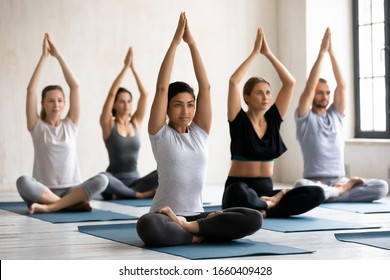 Attractive Indian Ethnicity Female Coach Lead Yoga Class Teach Caucasian Group Of People Meditating Together, Girls And Guys Raised Arms Up Make Namaste Gesture Seated In Lotus Pose Working Out Indoor