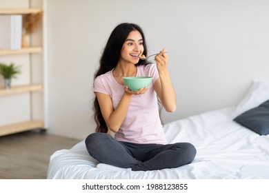 Attractive Indian lady eating yummy cereal with milk while sitting on comfortable bed at home, full length. Young Eastern woman having wholesome meal, keeping balanced weight loss diet