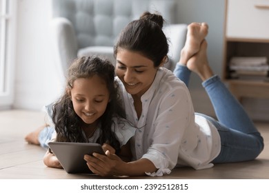 Attractive Indian preschool daughter young mother lying on warm floor in living room using digital tablet, play on-line video games, spend time on internet at home. Modern technology, leisure concept - Powered by Shutterstock