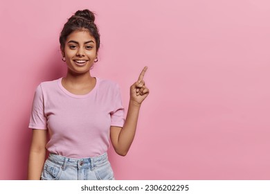 Attractive Indian teenage girl with dark hair points index finger above on copy space shows promotion offer demonstrates advertisement dressed in casual t shirt and jeans isolated over pink background - Powered by Shutterstock