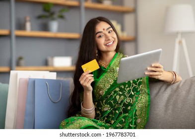 Attractive Indian woman in ethnic sari dress using credit card and tablet computer for online shopping at home. Pretty millennial lady buying goods on web, surrounded by gift bags