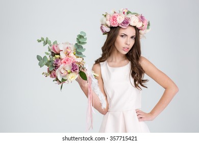 Attractive irritated young woman in white dress and wreath of roses showing bouquet of flowers over white background