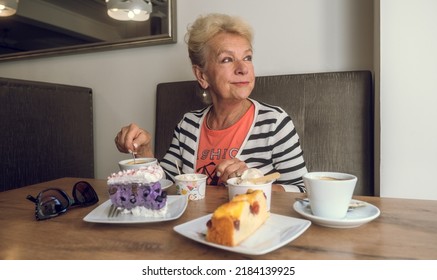 Attractive, Joyful Older Woman Drinking Coffee While Sitting At A Cafe. Portrait Of Mature Woman In A Cafeteria. Colorful Cakes And Coffee On The Table. Pleasant Relaxing Atmosphere.
