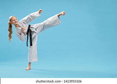 Attractive karate girl practicing kick foot forward. Cheerful, pretty child in white kimono with black belt , barefood posing in studio on blue background. Positive kid in pose of karate.