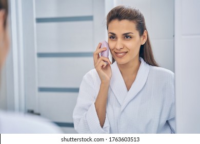 Attractive Lady In Bathrobe Using Facial Cleaning Massage Brush And Smiling