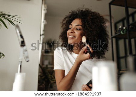 Attractive lady contouring face. Dark-skinned woman holds makeup brush, powders, looks into mirror at home.