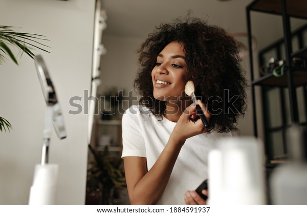 Attractive lady\
contouring face. Dark-skinned woman holds makeup brush, powders,\
looks into mirror at\
home.