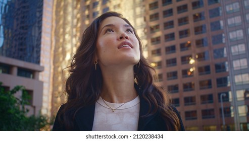 Attractive lady looking around whirling on modern street closeup. Cute brunette girl exploring megapolis on summer weekend. Young asian tourist watching high beautiful skyscraper. Happy active leisure