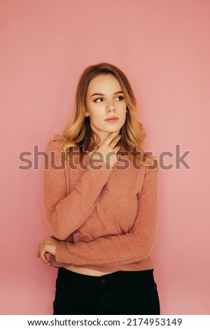 Attractive lady in a pink sweater posing for the camera on a pink background, looking away with a serious face. Vertical.