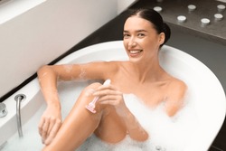 Attractive Lady Shaving Legs Smiling To Camera Using Pink Safety Razor Posing In Bathtub Full Of Foam Bubbles In Modern Bathroom Indoor. Female Enjoying Hair Removal Routine Taking Bath