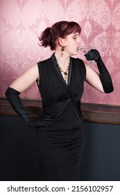 attractive lass wearing black frock and long black gloves is standing and having strong drinks