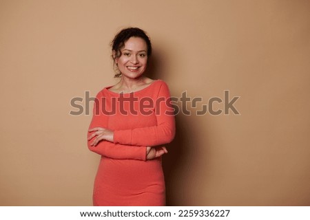 Attractive Latin American middle-aged woman, wearing bright orange casual dress, smiling a beautiful toothy smile looking at camera, posing with her arms crossed over beige background. People concept