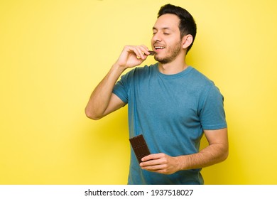 Attractive Latin Man Eating A Big Chocolate Candy Bar. Handsome Man In His 30s With Sugar Cravings Enjoying A Chocolate