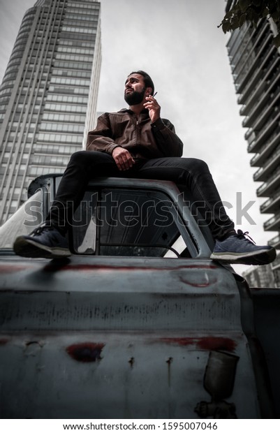 Attractive latin model, handsome brunet men\
with beard, cigarette, leather jacket sits on a roof of  car,\
smokes in the city center of Buenos Aires. Urban, seriously male\
portrait and modern\
buildings