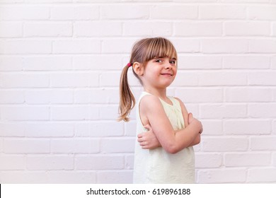 Attractive Little Blonde Girl Child Profile Half Length Portrait. Cute Kid With Arms Folded Against White Brick Wall