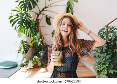 Attractive Long-haired Girl In Hat With Nude Makeup Standing With Cocktail In Room With Big Plants In Pots. Indoor Portrait Of Glad Young Woman In Wristwatch Holding Glass Of Juice.